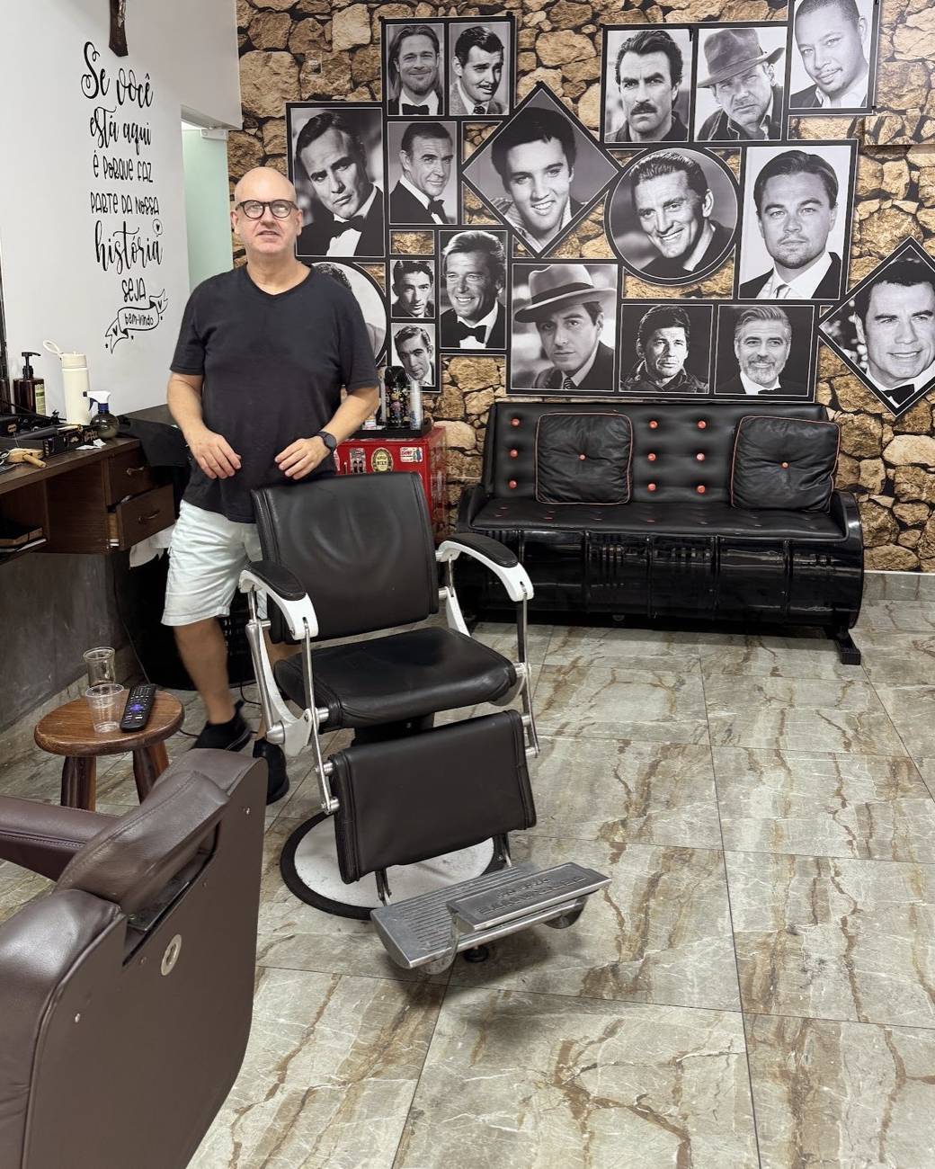 Ed atrás da cadeira de barbeiro, com a parede de pedra e quadros ao fundo